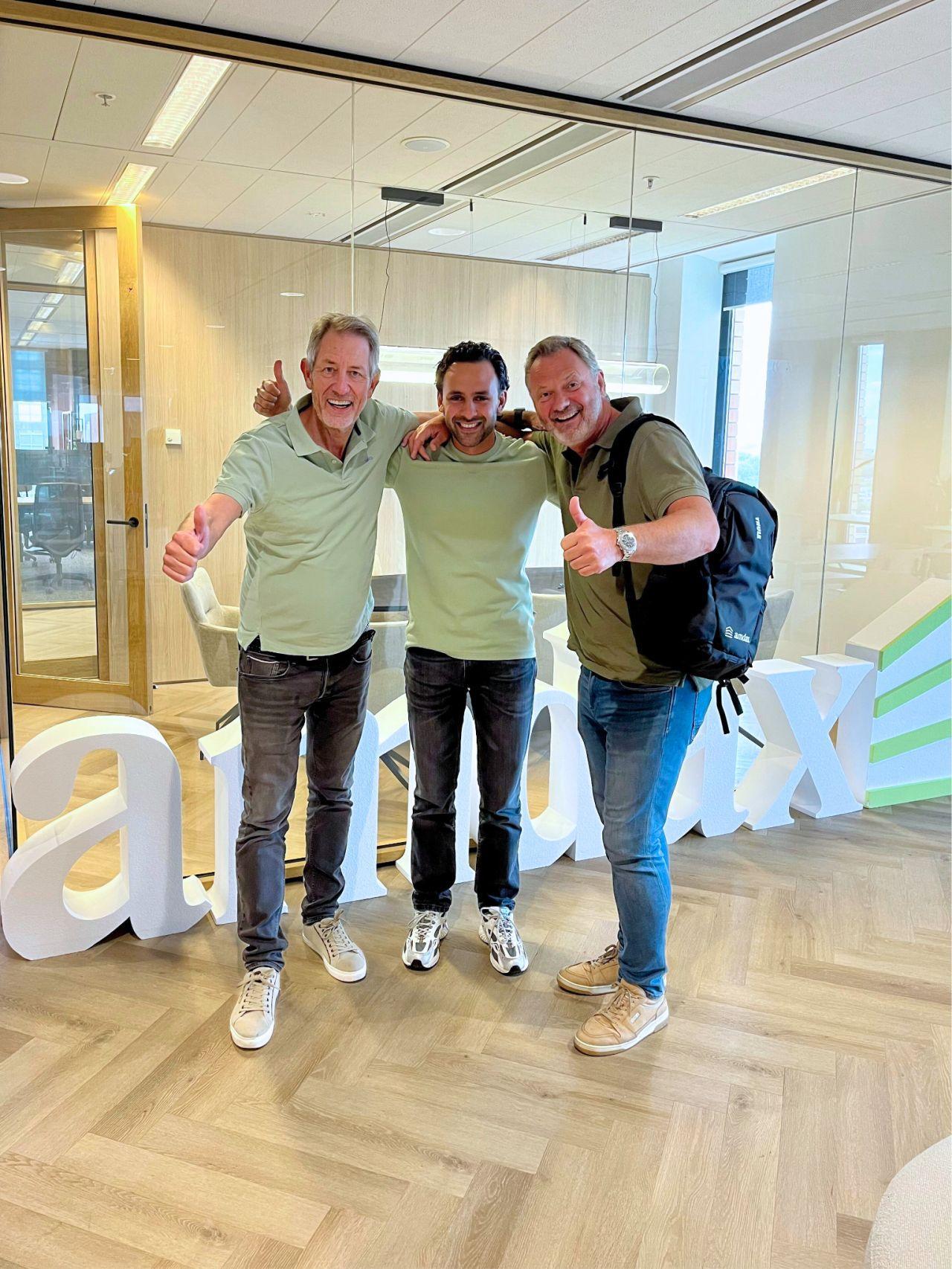 Three men giving thumbs up in front of large white Amdax letters in an office